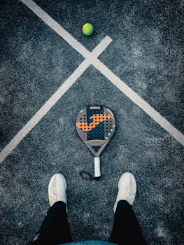 Flat lay of a padel racket, ball, and shoes on textured court surface.